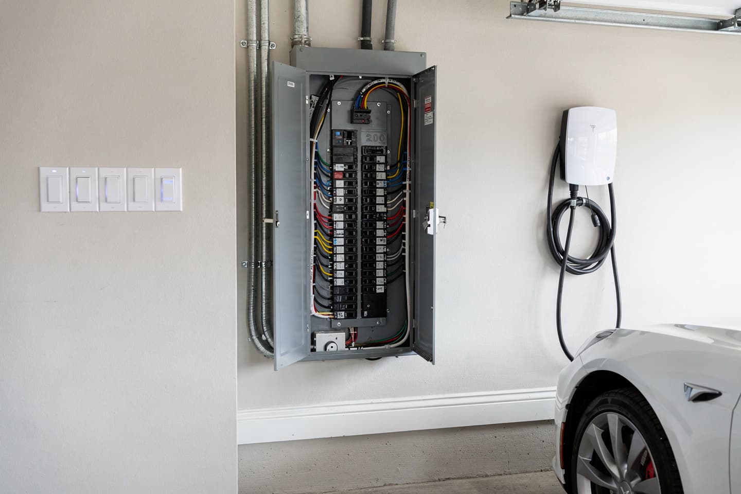 Open 200-amp breaker panel with labeled circuits in a garage beside a wall-mounted Level 2 EV charger and a row of smart switches