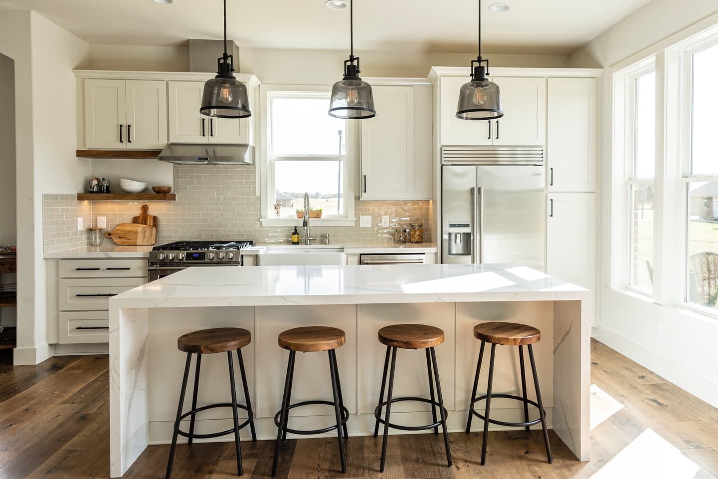 Remodeled kitchen with white shaker cabinets, a quartz waterfall island with four wood-top stools, black and smoked-glass pendant lights, a gray subway tile backsplash, a stainless range and refrigerator, and wide-plank wood floors